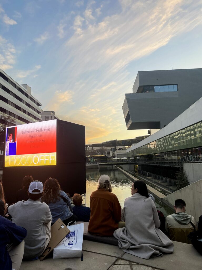 Photo of sun setting over a brutalist architecture with people watching a open-air presentation.